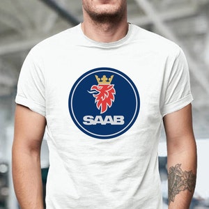 May include: White t-shirt featuring the Saab logo. The logo is a blue circle with a red griffin and a gold crown, and the word "SAAB" in white. The shirt is short-sleeved and worn by a person.