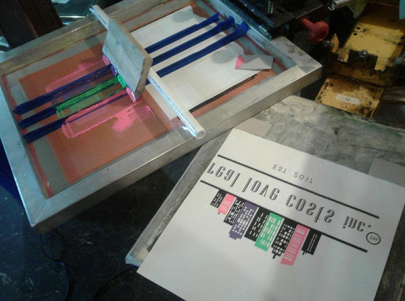 Pode incluir: Uma configura&ccedil;&atilde;o de serigrafia com uma moldura de serigrafia e uma folha de papel por baixo. A moldura de serigrafia tem tinta azul, verde e rosa. O papel tem um desenho em preto e branco de um horizonte da cidade com o texto "mc. 212 CO2/G 10AG 1691" e "T105 E21".