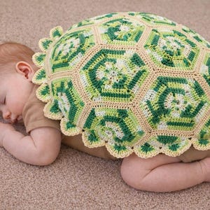 May include: A sleeping baby is nestled under a crocheted turtle shell blanket. The blanket features a green, white, and tan geometric pattern with a scalloped edge. The baby is wearing a brown onesie and is lying on a neutral-colored carpet.