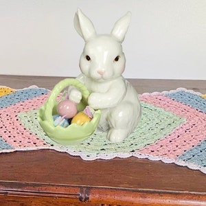 May include: A white ceramic bunny figurine holding a green basket filled with colorful Easter eggs. The bunny is sitting on a multi-colored crocheted doily.