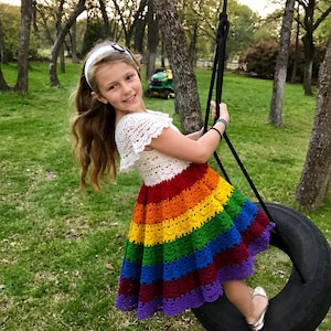 Könnte beinhalten: Ein handgehäkeltes Kleid mit weißem Kurzarm-Oberteil und einem ausgestellten Rock mit horizontalen Streifen in Rot, Orange, Gelb, Grün, Blau und Violett. Das Kleid wird mit einem weißen Stirnband und silberfarbenen Ballerinas kombiniert.