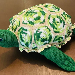 May include: A crocheted green and beige turtle with a detailed shell pattern. The turtle is lying on a wooden surface.