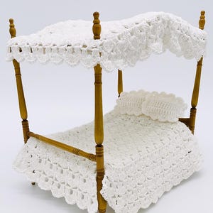 May include: A miniature four-poster bed with a white crocheted canopy, blanket, and pillow. The bed frame is made of light brown wood. The canopy and blanket have a scalloped edge design. The bed is a decorative item.