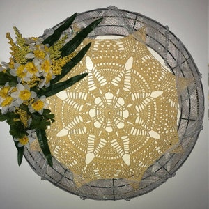 May include: A crocheted doily with a delicate floral design in a beige color. The doily is displayed on a silver wire wreath with white beads. A bouquet of yellow daffodils is placed on the wreath.
