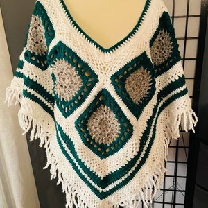 May include: A white crochet poncho with green and grey accents. The poncho has a v-neck and fringe trim.