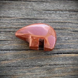 Puede incluir: Una talla de piedra pulida de color marrón rojizo con forma de oso de perfil. La figura presenta una superficie lisa y brillante con el lomo redondeado y una sección recortada distintiva entre las patas delanteras y traseras.