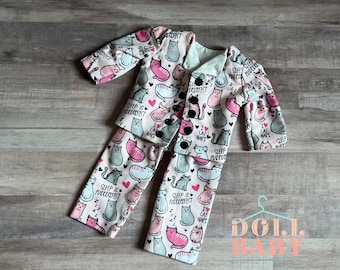 Puede incluir: Un pijama rosa y azul para una muñeca con estampado de gato. El pijama tiene una chaqueta con botones y pantalones. El estampado presenta gatos, corazones y el texto "Sleep is Purrfect" y "I love cat naps".