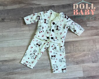 Puede incluir: Un conjunto de pijama blanco con una parte superior abotonada y pantalones. La pijama presenta un estampado de animales de dibujos animados con botones negros y detalles rojos. El texto "Cute as a button" está impreso en los pantalones.