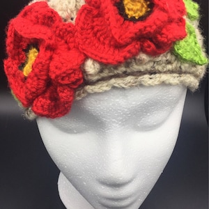 May include: A crocheted headband with two red poppy flowers and green leaves. The headband is beige and has a textured pattern.