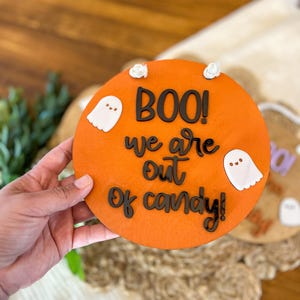 以下が含まれることがあります： オレンジ色の丸い木製看板に、「BOO! We are out of candy!」という黒い文字が書かれています。看板には白い幽霊の切り抜きが2つあります。