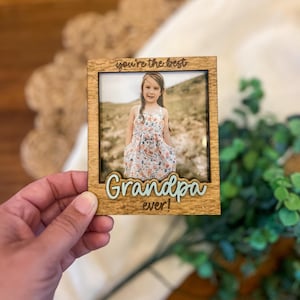 Op de afbeelding: Een houten fotolijst met een foto van een jong meisje in een bloemenjurk. De lijst heeft de tekst "you're the best Grandpa ever!" in een lichtblauwe lettertype.