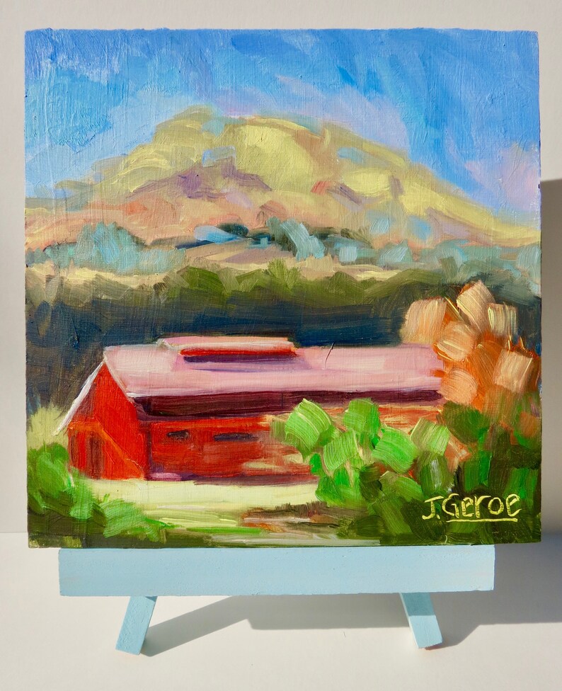 May include: A painting of a red barn with a green bush in the foreground. The barn is set against a backdrop of a rolling green hill with a yellow-green mountain in the distance. The sky is blue with white clouds. The painting is signed "J. Geroe" in the lower right corner.