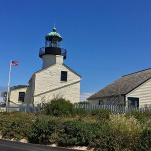 May include: A white lighthouse with a green roof and a black railing. The lighthouse is on a hill with a white picket fence and green bushes. An American flag is flying in front of the lighthouse.