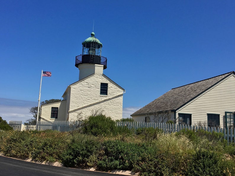 May include: A white lighthouse with a green roof and a black railing. The lighthouse is on a hill with a white picket fence and green bushes. An American flag is flying in front of the lighthouse.