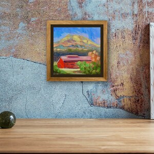 May include: A framed painting of a red barn with a green roof, surrounded by trees and a hill in the background. The sky is blue with white clouds. The painting is in a brown frame.