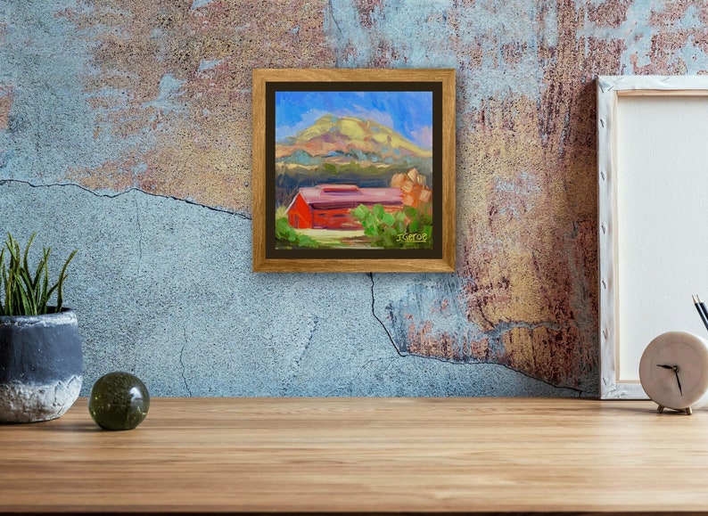 May include: A framed painting of a red barn with a green roof, surrounded by trees and a hill in the background. The sky is blue with white clouds. The painting is in a brown frame.
