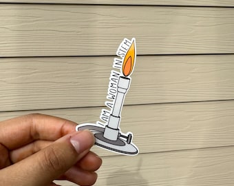 May include: A white sticker with a drawing of a Bunsen burner with a flame on top. The sticker has the text "I AM A WOMAN IN STEM" written vertically on the side of the burner.