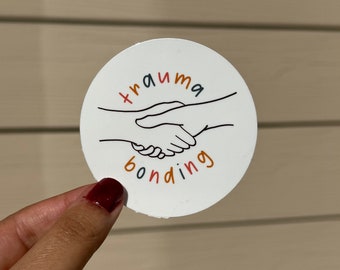 May include: White round sticker with a black line drawing of two hands shaking. The text "trauma bonding" is written in colorful letters below the hands.