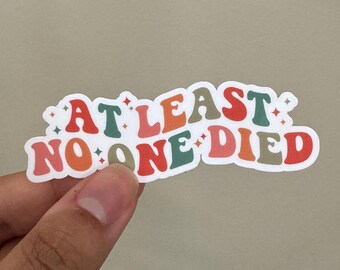 May include: A white sticker with the text "AT LEAST NO ONE DIED" in colorful, retro-style lettering. The letters are outlined in black and have a slight shadow effect. The sticker has a glossy finish.