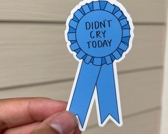 May include: A blue ribbon sticker with the text "Didn't Cry Today" in black lettering. The ribbon is tied in a bow at the top.