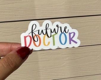 May include: A white sticker with the words "future DOCTOR" in a rainbow of colors. The word "future" is in black cursive, and "DOCTOR" is in a bold, colorful font. The sticker has a cloud-like shape.