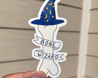 May include: A white bone with a blue wizard hat with yellow stars and crescent moons. The bone has a banner that says "BONE WIZARD".