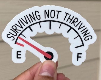May include: White sticker with a black and white gas gauge design. The gauge reads "SURVIVING NOT THRIVING" with a red needle pointing to "E".