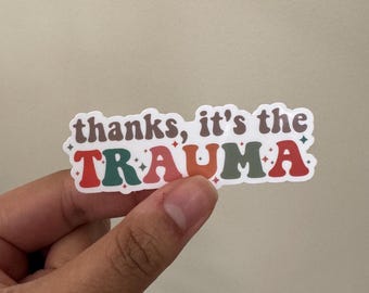 May include: A white sticker with the text "thanks, it's the trauma" in colorful, retro-style lettering with a glittery effect.