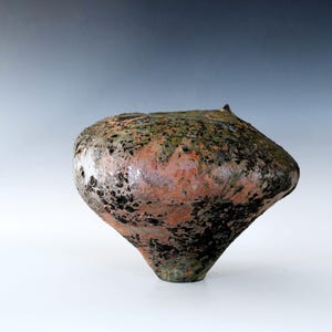 May include: A handcrafted ceramic vase with a unique, organic shape. The vase features a blend of colours, including shades of orange, green, and black, creating a textured, earthy appearance. The vase is approximately 25 cm tall.