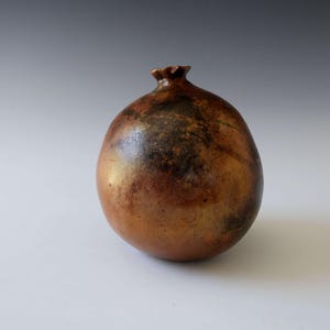 Puede incluir: Escultura de cerámica artesanal con forma de granada. La pieza, con forma de fruta, presenta un cuerpo liso y redondeado y una parte superior texturizada. El color es una mezcla de marrón, dorado y negro, con un acabado brillante. Objeto decorativo.
