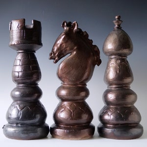 May include: Three bronze-colored chess pieces: a rook, a knight, and a bishop. The rook has a crenellated top, the knight features a horse head, and the bishop has a rounded top. Each piece has a textured surface and a dark metallic finish, suitable for display.