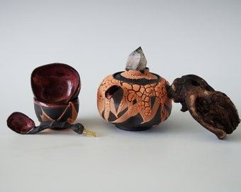 May include: A collection of handcrafted ceramic pieces. Includes a small teapot with a crystal, two stacked cups, and a spoon. The teapot has a cracked orange and black design. The spoon and cups are dark with red interiors.
