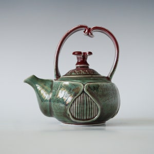 Può includere: Teiera in ceramica artigianale dal design unico. La teiera presenta un corpo verde con superficie strutturata e manico e coperchio bordeaux. Il manico è elegantemente curvo e il beccuccio è finemente modellato.