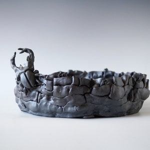 Op de afbeelding: Een handgemaakte, donkergrijze keramische kom met een getextureerde, steenachtige buitenkant. De kom heeft een abstracte, sculpturale handgreep aan één kant, wat een uniek artistiek element toevoegt. Geschikt voor decoratief gebruik.