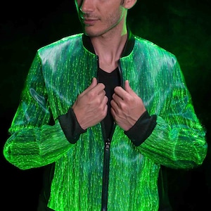 Pode incluir: Uma jaqueta bomber verde com fecho de correr e forro interior preto. A jaqueta é feita de um tecido que tem luzes de fibra óptica embutidas que criam um efeito brilhante. A jaqueta tem a palavra "YMPU" impressa na parte inferior.