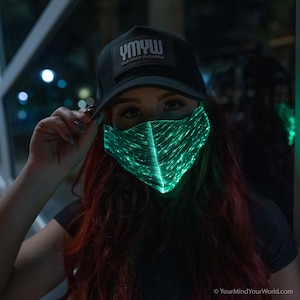 May include: A person wearing a black cap with the text "YMYW Your Mind Your World" and a green LED light-up face mask.
