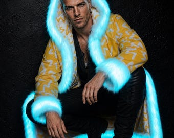 May include: A long coat featuring a yellow fabric with a white zigzag pattern, accented by a bright cyan faux fur trim along the hood, cuffs, and hem. The coat is worn over a black shirt and black pants, creating a high-contrast visual appearance.