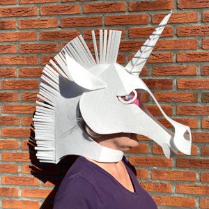 May include: A white unicorn mask with a silver horn and pink eye detail. The mask has a mane of white fringe and is worn by a person wearing a purple shirt. The background is a red brick wall.