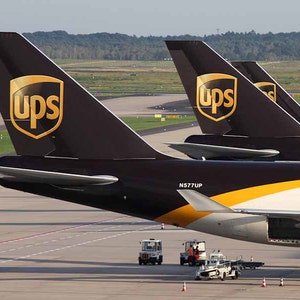Peut inclure: Gros plan sur les empennages de plusieurs avions cargo UPS. Les empennages sont brun fonc&eacute; avec un logo UPS dor&eacute;. Les avions sont stationn&eacute;s sur un tarmac avec des v&eacute;hicules de service &agrave; proximit&eacute;.