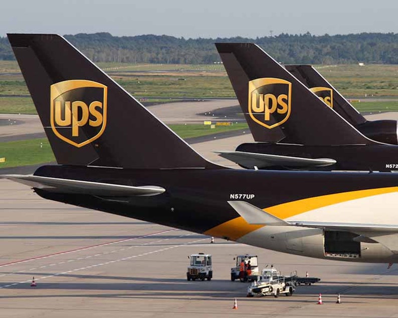 Peut inclure: Gros plan sur les empennages de plusieurs avions cargo UPS. Les empennages sont brun fonc&eacute; avec un logo UPS dor&eacute;. Les avions sont stationn&eacute;s sur un tarmac avec des v&eacute;hicules de service &agrave; proximit&eacute;.