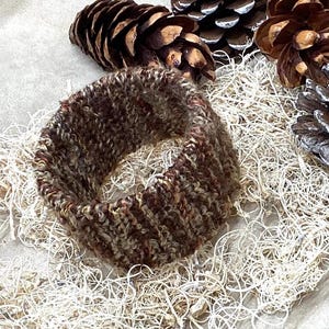 Könnte beinhalten: Ein braunes und beigefarbenes Strickstirnband. Das kreisförmige Stirnband besteht aus strukturiertem Garn. Der Hintergrund umfasst Tannenzapfen und getrocknetes Pflanzenmaterial, wodurch eine natürliche, erdige Ästhetik entsteht. Das Stirnband ist ein gemütliches Accessoire.