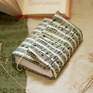 May include: A book wrapped in a fabric cover printed with musical notes, tied with twine. The book is on a neutral-toned surface, with a green fabric and dried plants in the background. An open book is visible in the background.
