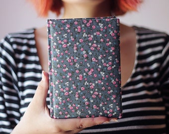 Puede incluir: Un libro rectangular con una cubierta estampada con flores. La cubierta es de color gris oscuro con un patrón de rosas rosas, pequeñas flores blancas y hojas azules. El libro se sostiene frente a una persona que lleva una camisa a rayas blancas y negras.