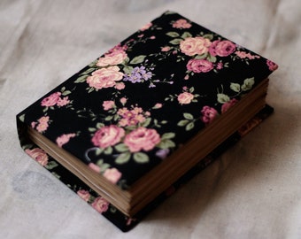 Puede incluir: Un libro de tapa dura con una cubierta de tela floral negra con rosas rosas y melocotón, hojas verdes y acentos morados. Las páginas del libro son visibles y la estética general es vintage.
