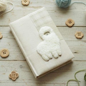 May include: A cream-coloured journal with a fluffy white alpaca design is surrounded by balls of yarn in cream, blue, and brown, along with wooden buttons. The items are arranged on a light-coloured wooden surface.