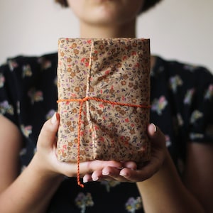 May include: A wrapped gift with a floral pattern in shades of brown, red, and purple. The gift is tied with orange twine. The person holding the gift is wearing a black floral top.