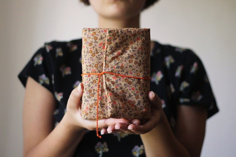 May include: A wrapped gift with a floral pattern in shades of brown, red, and purple. The gift is tied with orange twine. The person holding the gift is wearing a black floral top.