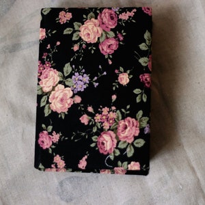 Puede incluir: Una cubierta de libro rectangular negra con un estampado floral. El dise&ntilde;o presenta rosas rosas y melocot&oacute;n, flores moradas y hojas verdes. La cubierta se encuentra sobre un fondo neutro y texturizado.