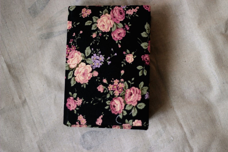 Puede incluir: Una cubierta de libro rectangular negra con un estampado floral. El dise&ntilde;o presenta rosas rosas y melocot&oacute;n, flores moradas y hojas verdes. La cubierta se encuentra sobre un fondo neutro y texturizado.