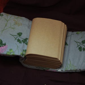 May include: An open journal with blank pages rests on a floral patterned fabric. The fabric is light gray with green and pink floral designs. The journal's pages are a light brown color.